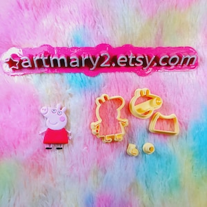 Puede incluir: Un juego de cortadores de galletas de plástico amarillo con un cerdo de dibujos animados, un vestido, un sombrero y un par de zapatos. Los cortadores están sobre una superficie peluda rosa y azul. El texto "artmary2.etsy.com" es visible en la imagen.