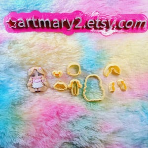 May include: A set of yellow plastic cookie cutters featuring a girl with long blonde hair, a dress, a hat, and other clothing items. The cutters are on a pink and blue fuzzy surface. The text "artmary2.etsy.com" is visible at the top of the image.