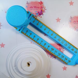 May include: A blue plastic circle cutter with a white plastic base and a gold ruler marked with numbers from 3 to 15. The cutter is on a white surface with pink stars and a pink and white floral design with the text "Art Mary".