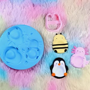 Peut inclure: Un moule en silicone bleu rond avec trois cavités, un emporte-pièce en plastique rose et trois figurines finies : une abeille jaune et noire, un pingouin noir et blanc, et un axolotl rose clair avec un visage souriant et de petits membres.
