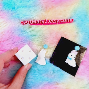 May include: White bunny rabbit charm with a blue bow and a blue balloon. The charm is on a white display stand with four holes. The charm is also shown on a black background.