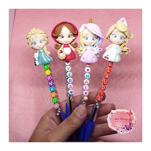 May include: Four decorative pens with character figurines. Each pen features a different character: a princess, Little Red Riding Hood, a unicorn, and a nurse. The names "Fernanda", "Vera", "Maea", and "Claudia" are spelled out with colorful beads on each pen.