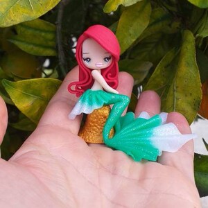 May include: A small, red-haired mermaid figurine with a green and white tail. The figurine is sitting on a gold base and is holding a seashell.