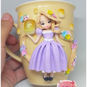 May include: A ceramic mug with a hand-painted design featuring a Rapunzel doll holding a lantern. The doll is wearing a purple dress and has long blonde hair. The mug is decorated with flowers and vines.