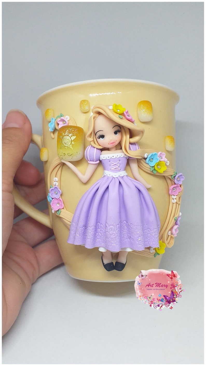 May include: A ceramic mug with a hand-painted design featuring a Rapunzel doll holding a lantern. The doll is wearing a purple dress and has long blonde hair. The mug is decorated with flowers and vines.