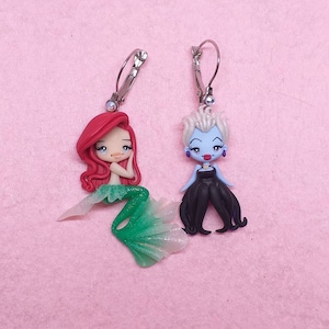 May include: A pair of clip-on earrings featuring a cartoon mermaid and Ursula from Disney's The Little Mermaid. The mermaid is wearing a green tail and has red hair. Ursula is wearing a black dress and has purple hair.