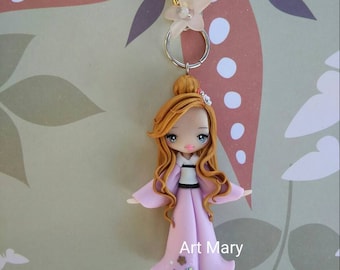 Op de afbeelding: Een roze en witte pop met lang bruin haar, die een roze kimono draagt met bloemmotieven. De pop is bevestigd aan een gouden ketting met een transparante bloemenhanger en een zilveren ring.
