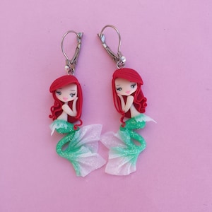 May include: A pair of earrings featuring a mermaid with red hair, a green tail with glitter, and a white iridescent tail fin. The mermaid is wearing a silver earring.