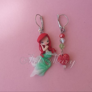 May include: A pair of earrings featuring a red-haired mermaid charm with a green tail and a red heart charm with clear crystal beads.