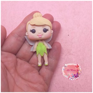 Peut inclure: Une petite figurine peinte à la main de la fée Clochette, avec des cheveux blonds, une robe verte et des ailes blanches. La figurine est en pâte polymère et a un style cartoon.