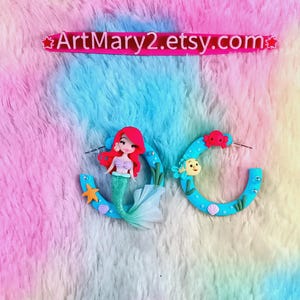 May include: A pair of blue hoop earrings with mermaid and sea creature designs. One earring features a mermaid with red hair and a green tail. The other earring has a yellow fish and a red crab. The text "ArtMary2.etsy.com" is above the earrings.