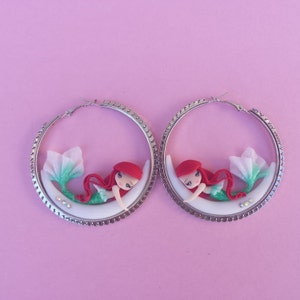 May include: A pair of silver hoop earrings with a white crescent moon and a mermaid figurine. The mermaid has red hair, a green tail, and a white and green top.