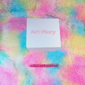 May include: White box with the text "Art Mary" printed in pink on the front. The box is sitting on a colorful fluffy surface.