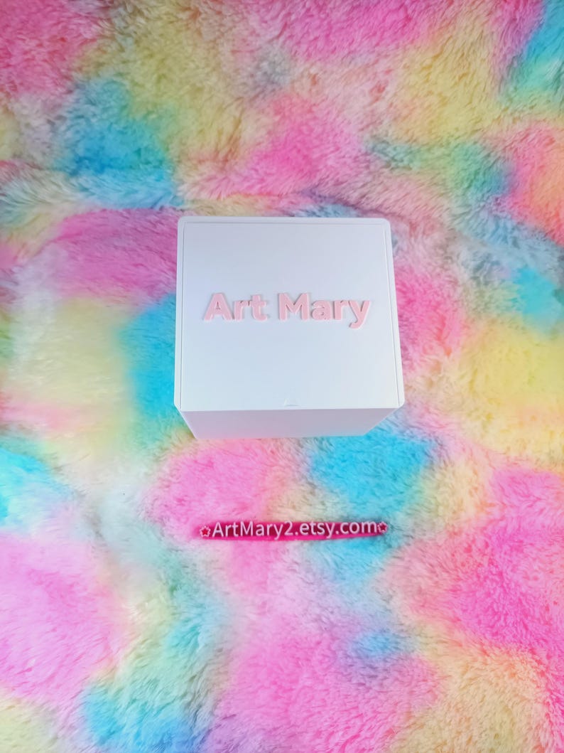 May include: White box with the text "Art Mary" printed in pink on the front. The box is sitting on a colorful fluffy surface.