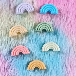 May include: Seven small, semi-circular rainbow-shaped charms in pastel colors including lavender, blue, peach, mint green, pink, white, and yellow. Each charm features three concentric arched ridges and a flat base, arranged in a scattered pattern.