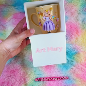 May include: A white box with a yellow mug inside. The mug has a floral design and a small figurine of a girl with blonde hair and a purple dress. The box has the text "Art Mary" on the front and the text "ArtMary2.etsy.com" on the bottom.
