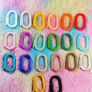 Puede incluir: Una colección de veinte mosquetones de forma ovalada dispuestos en filas. Los clips están hechos de plástico y vienen en una variedad de colores sólidos que incluyen blanco, amarillo, naranja, rojo, rosa, morado, verde, azul, marrón, beige y negro.