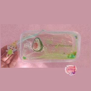 Peut inclure: Une pochette en plastique transparente avec un personnage d'avocat vert et le texte "Cute Avocado" à l'intérieur. La pochette est recouverte de paillettes vertes et d'un petit charme de fée.
