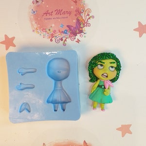 Puede incluir: Un molde de silicona para hacer una pequeña figura de muñeca. El molde tiene tres secciones: un cuerpo, un par de brazos y un par de piernas. El molde es azul y tiene un fondo blanco. Hay una pequeña figura de muñeca verde y rosa al lado del molde.