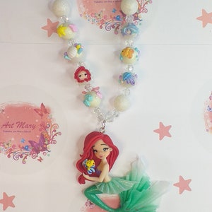 May include: A handmade necklace with a mermaid charm. The necklace features a variety of beads, including clear, white, and glitter beads. The mermaid charm is made of polymer clay and is painted in shades of red, green, and yellow. The necklace is a great gift for any little girl who loves mermaids.