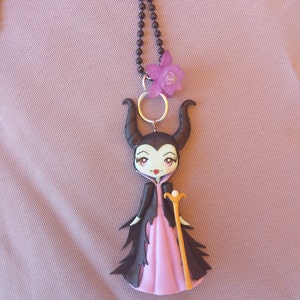 May include: A black necklace with a purple flower charm and a pendant of a cartoon Maleficent from Disney's Sleeping Beauty. The pendant is made of polymer clay and features Maleficent in a pink dress and black cape, holding a golden staff.