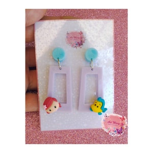 May include: A pair of lavender dangle earrings with a light blue round charm at the top. One earring features a small red and yellow figurine of Ariel from The Little Mermaid. The other earring features a small yellow and green figurine of Ursula from The Little Mermaid. The earrings are displayed on a white card with the text "Art Mary" printed on it.