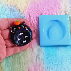 Pode incluir: Um prato decorativo preto brilhante em forma de gato com uma pequena abóbora laranja na cabeça e padrões de espinhas de peixe brancas, posicionado ao lado de um molde de silicone azul claro quadrado com uma cavidade circular.