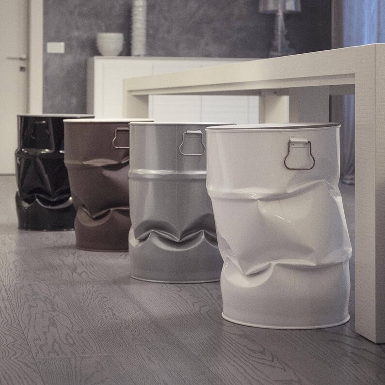 May include: Four metal barrel-shaped stools in a row, each with a unique color and dented design. Colors include black, brown, gray, and white. The stools have a handle on the top and sit on a wooden floor.