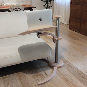 May include: A wooden laptop stand with a silver metal pole and a curved base. The stand is positioned next to a white sofa with a green cushion.