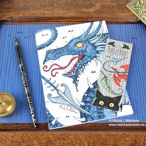 Blue Dragon Notebook featuring my original pen and ink dragon illustration, front cover, shown with bookmark and vintage Victorian writing desk accessories. Made in the UK. Design and illustration © Nicola L Robinson | Teeth and Claws