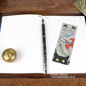 Blue Dragon Notebook, featuring my original pen and ink dragon illustration, open flat showing lined pages, with bookmark and vintage Victorian writing desk accessories. Made in the UK. Design and illustration © Nicola L Robinson | Teeth and Claws
