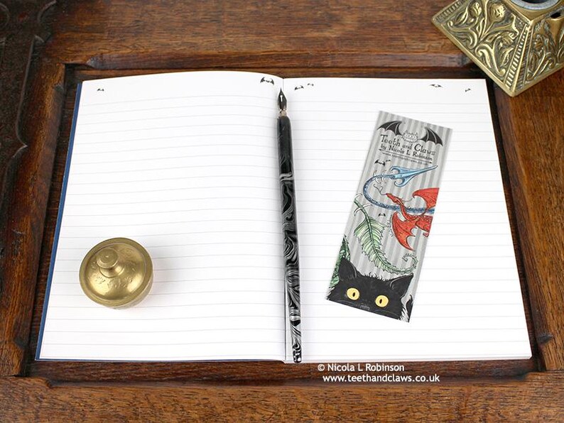 Blue Dragon Notebook, featuring my original pen and ink dragon illustration, open flat showing lined pages, with bookmark and vintage Victorian writing desk accessories. Made in the UK. Design and illustration © Nicola L Robinson | Teeth and Claws