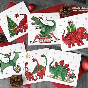 Może przedstawiać: Zestaw kartek świątecznych z ilustracjami dinozaurów. Każda kartka przedstawia innego dinozaura z elementami świątecznymi, takimi jak czapki Mikołaja i prezenty. Kartki są na czerwonych kopertach, ze śnieżynkami i gwiazdką.