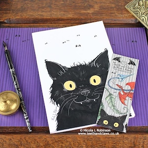 Może przedstawiać: Ilustracja czarnego kota z żółtymi oczami i kłami na białej karcie. Zakładka z napisem "Teeth and Claws" oraz pióro ze złotym kałamarzem leżą na fioletowej powierzchni. Widoczny jest adres strony internetowej.
