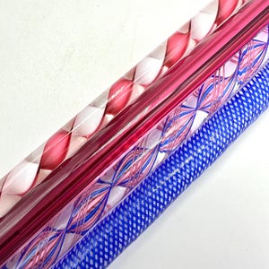 May include: Close-up of four glass rods with decorative patterns. The rods are in shades of pink and blue, with spiral, crisscross, and lattice designs. The rods are angled diagonally against a white background.
