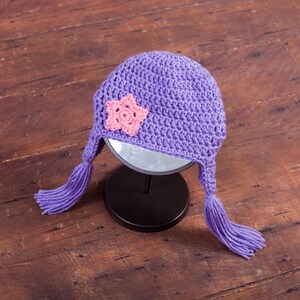May include: Purple crocheted hat with a pink flower embellishment and long tassels. The hat is sitting on a black stand.