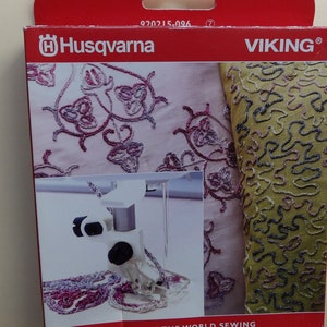 May include: A close-up of a white sewing machine foot attachment with a black knob. The foot is attached to a sewing machine needle and is positioned over a piece of fabric with intricate embroidery. The fabric is a light pink and green color with a floral design. The packaging is red and features the Husqvarna Viking logo and the text "Keeping the world sewing."