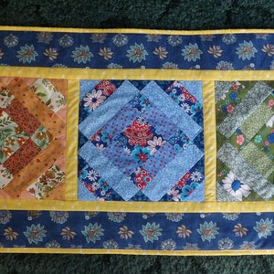 May include: A blue and yellow quilted table runner with three square panels featuring different floral patterns. The panels are surrounded by a yellow border and the runner has a blue floral border.