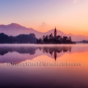 May include: A picturesque view of a church on an island in a lake at sunrise. The sky is a vibrant mix of pink, orange, and purple, reflecting in the still water. The mountains in the background are shrouded in mist.