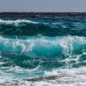 Può includere: Un'onda imponente si infrange sulla costa, creando uno spruzzo di schiuma bianca. L'acqua è di un blu turchese vibrante, a contrasto con il blu scuro dell'oceano sullo sfondo.