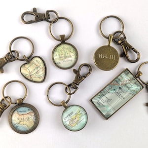 May include: A selection of antique brass keyrings with diverse map designs. The keyrings showcase round, heart-shaped, and rectangular pendants with detailed map graphics. Each keyring includes a clasp and a ring.