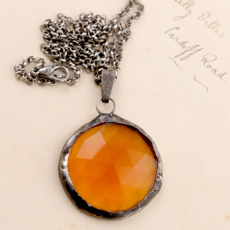 Puede incluir: Un collar de cadena de plata con un colgante de vidrio &aacute;mbar. El colgante es redondo y tiene un dise&ntilde;o facetado.