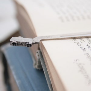 May include: A silver bookmark with a decorative design is placed in a book. The bookmark is shaped like a small, stylized figure with a rounded head and a long, thin body. The bookmark is placed in the book so that the head of the figure is visible and the body of the figure is hidden behind the pages of the book.