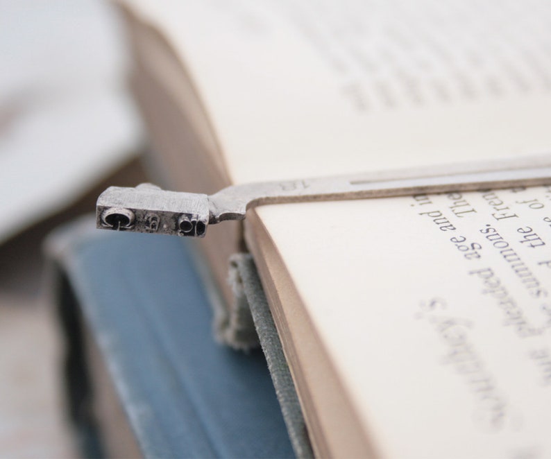 May include: A silver bookmark with a decorative design is placed in a book. The bookmark is shaped like a small, stylized figure with a rounded head and a long, thin body. The bookmark is placed in the book so that the head of the figure is visible and the body of the figure is hidden behind the pages of the book.