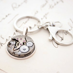 May include: A silver key chain with a round, open-faced watch charm. The watch face is exposed, revealing the inner workings of the watch mechanism. The watch face has a silver rim and a clear glass cover.