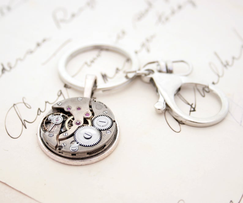 May include: A silver key chain with a round, open-faced watch charm. The watch face is exposed, revealing the inner workings of the watch mechanism. The watch face has a silver rim and a clear glass cover.