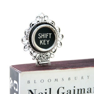May include: Silver bookmark featuring a black typewriter key that reads "SHIFT KEY". The bookmark is placed on a book with the title "Neil Gaiman" and "BLOOMSBURY" printed on the cover.