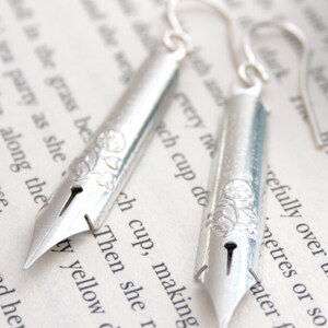 Puede incluir: Pendientes de plata con forma de plumines de caligraf&iacute;a vintage con detalles florales. Los pendientes cuelgan de una p&aacute;gina de libro con texto.