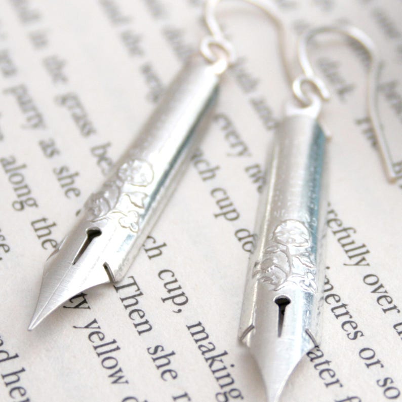 Puede incluir: Pendientes de plata con forma de plumines de caligraf&iacute;a vintage con detalles florales. Los pendientes cuelgan de una p&aacute;gina de libro con texto.