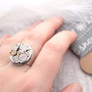 May include: A silver ring with a circular watch face design. The watch face is made of metal and has visible gears and a small pink jewel.